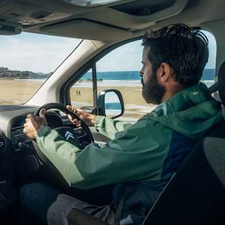 Man driving a van near a beach, holding the steering wheel, wearing a green jacket.