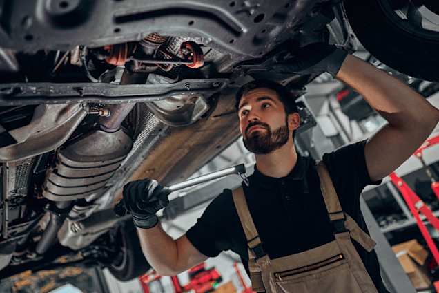 Mechanic inspecting a car's underside, holding a tool and wearing overalls.